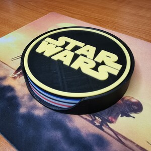 Puede incluir: Un posavasos redondo negro y amarillo con el texto "STAR WARS" en letras amarillas. El posavasos tiene una base negra con un dise&ntilde;o de rayas rojas, blancas y azules.