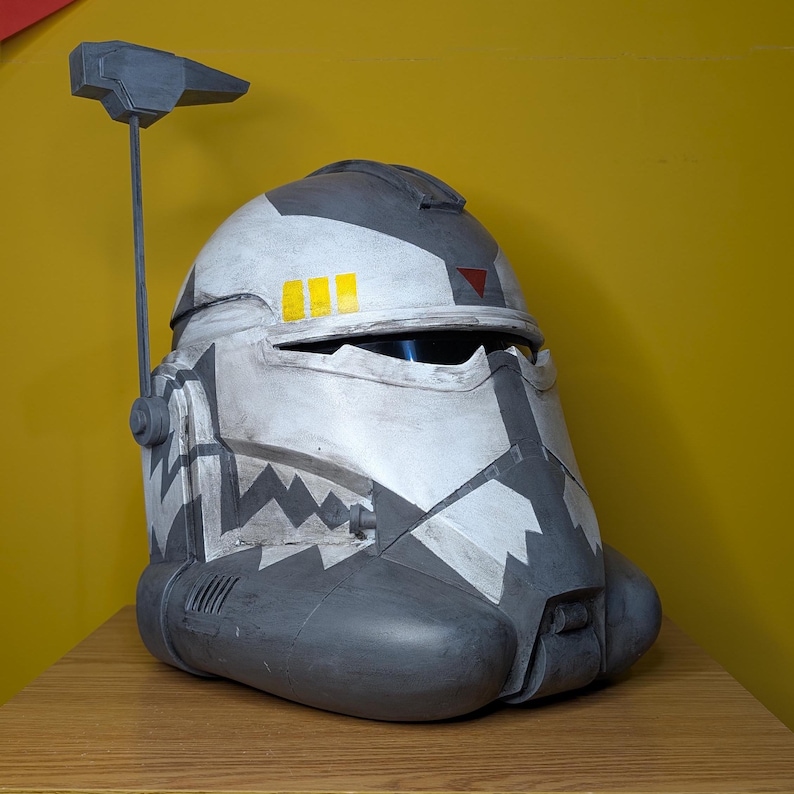 May include: A white and gray plastic helmet with a black visor and a gray antenna. The helmet has a black and white camouflage pattern and yellow and red markings.