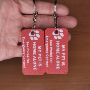 May include: Two red keychains with silver key rings. The keychains are rectangular and have white paw prints and the text "MY PET IS HOME ALONE" with instructions to see the wallet or purse for emergency contact.