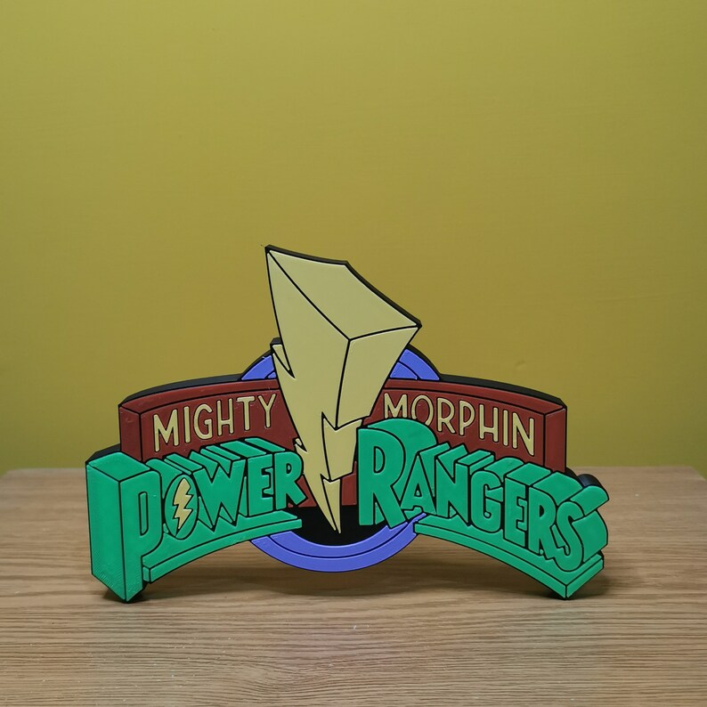 Pu&ograve; includere: Logo 3D dei Power Rangers in verde, rosso e giallo. Il logo mostra "Mighty Morphin Power Rangers" con un fulmine al centro. Il design ha un effetto ombra.
