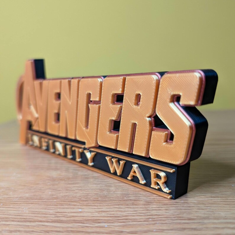May include: A 3D-printed sign featuring the word "AVENGERS" in orange, with "INFINITY WAR" below in white text on a black base. The sign has a layered design and is set against a light yellow background.
