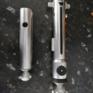 May include: Two silver lightsaber hilts, one is longer than the other. The longer hilt has a black section in the middle and a black cap on the bottom. The shorter hilt has a silver cap on the bottom.