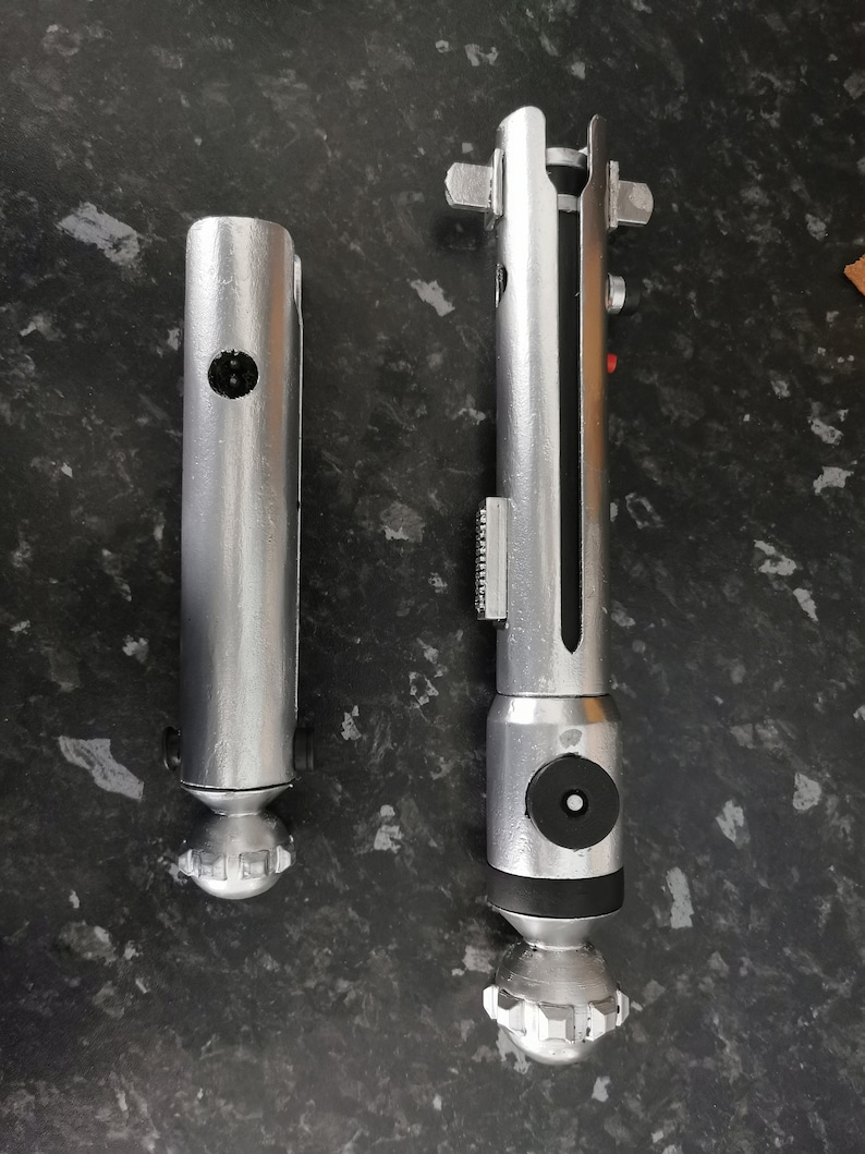 May include: Two silver lightsaber hilts, one is longer than the other. The longer hilt has a black section in the middle and a black cap on the bottom. The shorter hilt has a silver cap on the bottom.