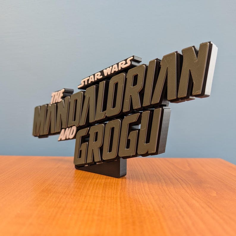 Op de afbeelding: Een 3D-geprint bord met de tekst "STAR WARS The Mandalorian and Grogu". Het bord heeft een zwarte basis en de tekst is in bruin- en wittinten. Het wordt weergegeven op een houten oppervlak.