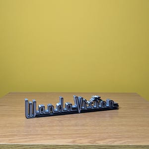 Peut inclure: Logo "WandaVision" noir et argent&eacute;, pos&eacute; sur une surface en bois devant un fond jaune. Le logo est un design stylis&eacute; en trois dimensions, avec un contour argent&eacute; m&eacute;tallique et un remplissage noir.
