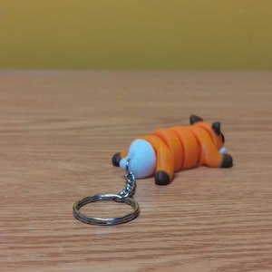 May include: Orange and white cartoon fox keychain with a silver key ring. The fox is lying on its back with its legs in the air.