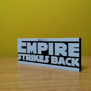 May include: A black and white sign with the text "THE EMPIRE STRIKES BACK" in white raised letters on a black background.