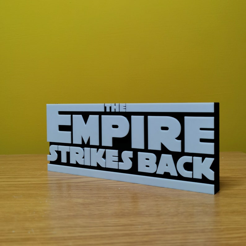 May include: A black and white sign with the text "THE EMPIRE STRIKES BACK" in white raised letters on a black background.