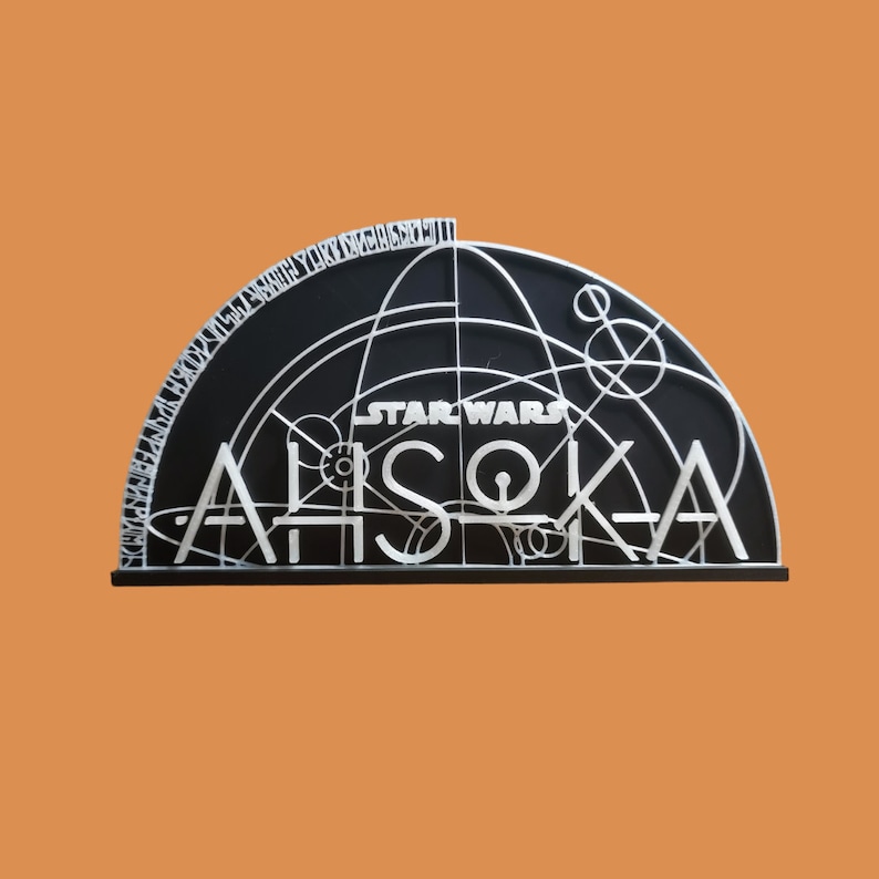 May include: A black and white 3D printed sign with the text "STAR WARS" and "AHsoka" in a stylized font. The sign features a geometric design with circles and lines.