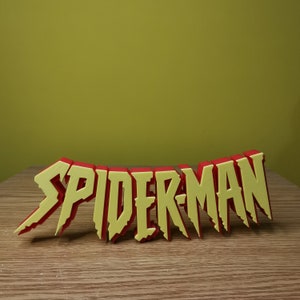 May include: A 3D printed word sculpture of "SPIDER-MAN" in red and yellow letters. The letters are stylized with jagged edges and a slightly distressed look.
