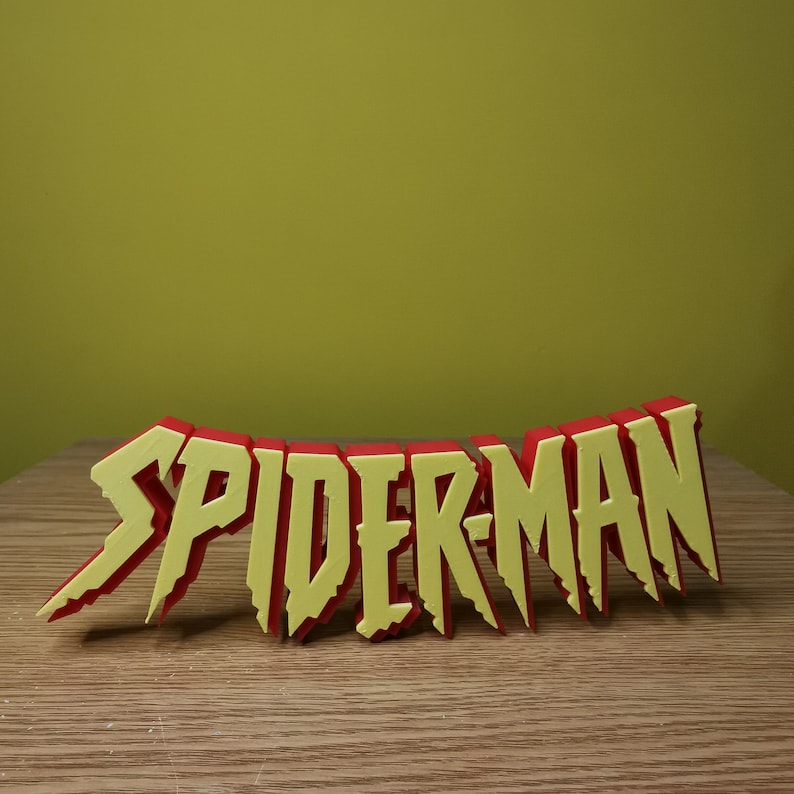May include: A 3D printed word sculpture of "SPIDER-MAN" in red and yellow letters. The letters are stylized with jagged edges and a slightly distressed look.