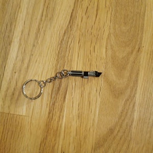 Op de afbeelding: Een zilveren sleutelhanger met een zwart en zilveren lightsaber-bedel. De lightsaber-bedel is ongeveer 5 cm lang.