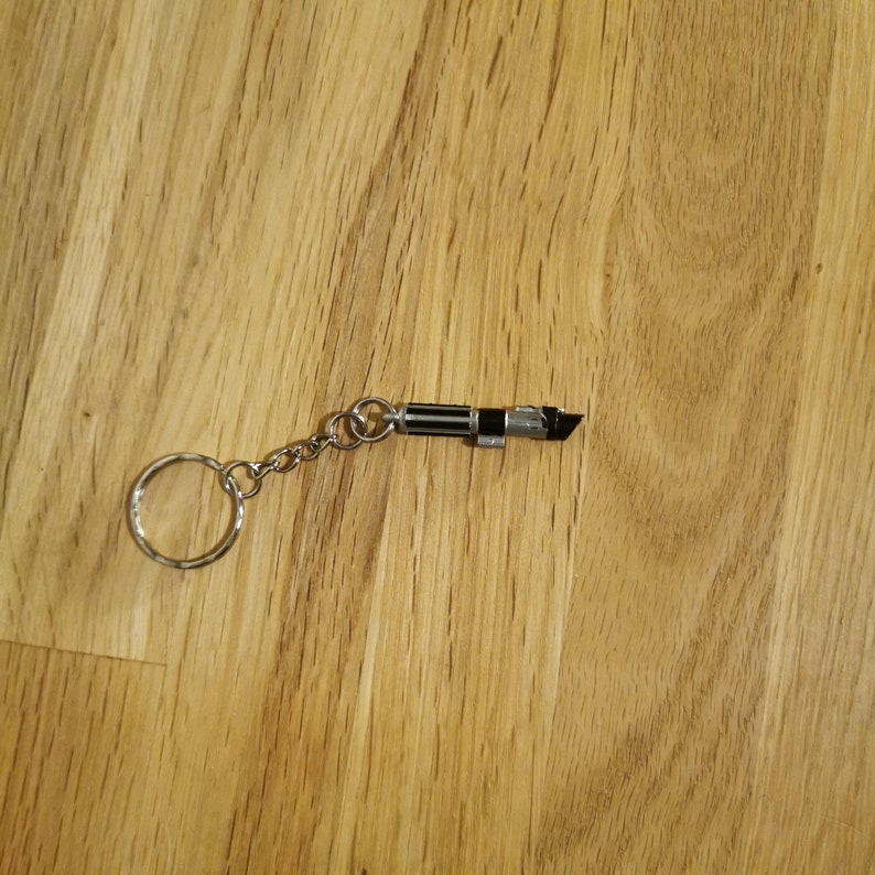 Op de afbeelding: Een zilveren sleutelhanger met een zwart en zilveren lightsaber-bedel. De lightsaber-bedel is ongeveer 5 cm lang.