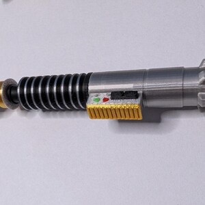 May include: A silver and black lightsaber replica with gold and yellow accents. The toy lightsaber has a green lanyard attached to the end. The lightsaber has a detailed design with buttons and a ribbed handle.