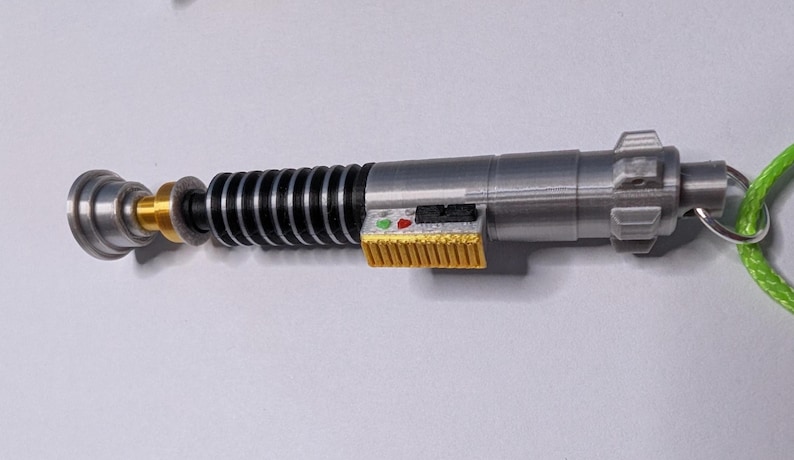 May include: A silver and black lightsaber replica with gold and yellow accents. The toy lightsaber has a green lanyard attached to the end. The lightsaber has a detailed design with buttons and a ribbed handle.