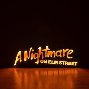 May include: Illuminated sign with the text "A Nightmare on Elm Street" in a stylized, orange font. The sign is lit from within, casting a warm glow against a dark background, with a reflection on the surface below.
