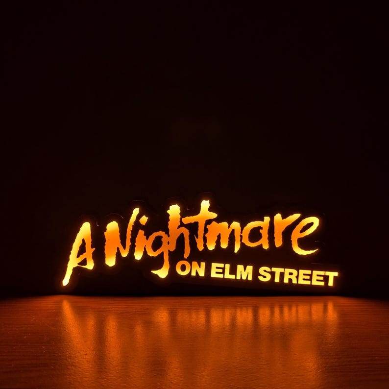 May include: Illuminated sign with the text "A Nightmare on Elm Street" in a stylized, orange font. The sign is lit from within, casting a warm glow against a dark background, with a reflection on the surface below.
