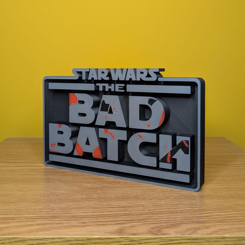 May include: A 3D printed sign with the text "STAR WARS THE BAD BATCH" in a black and gray color scheme. The text is in a bold, blocky font and has a distressed, weathered look. The sign is perfect for fans of the Star Wars: The Bad Batch animated series.