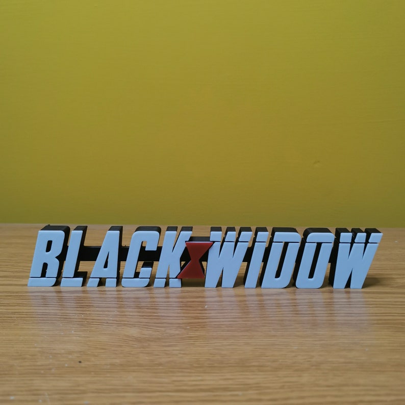 May include: White 3D letters spelling "BLACK WIDOW" with a red widow symbol in the middle. The letters are on a wooden surface.