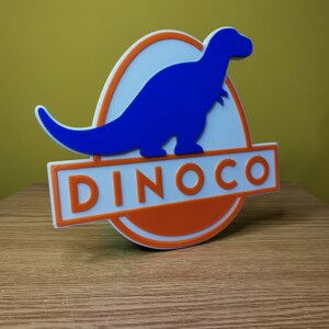 Puede incluir: Un letrero impreso en 3D con una silueta de dinosaurio azul y la palabra "DINOCO" en letras blancas sobre un fondo naranja.