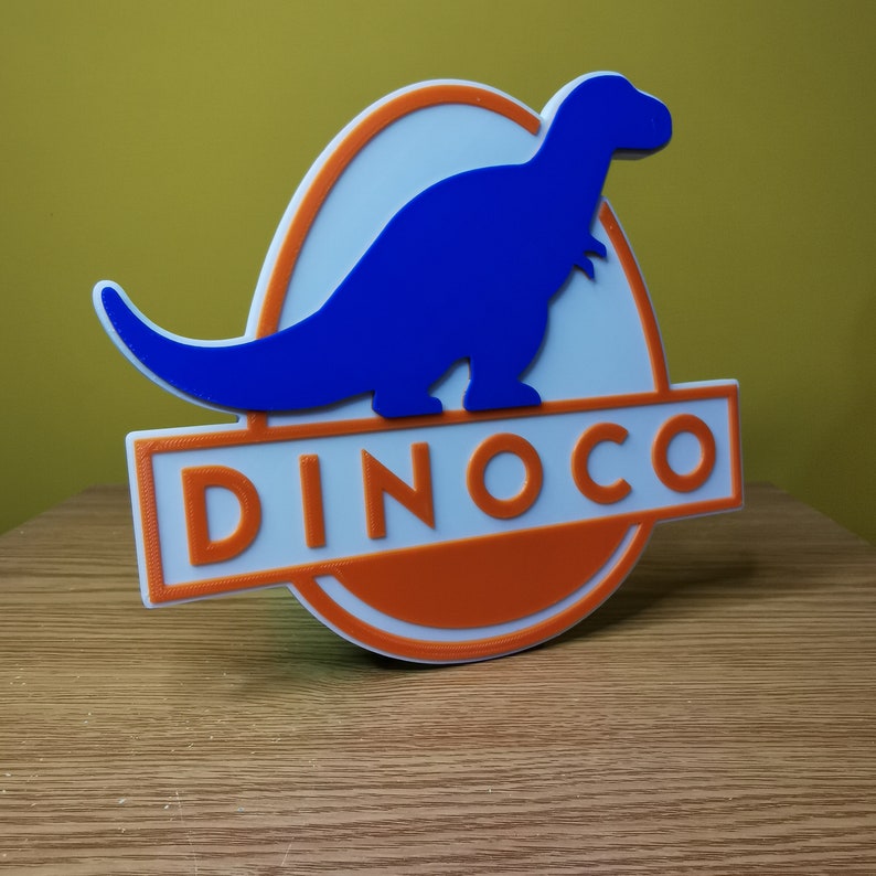 Puede incluir: Un letrero impreso en 3D con una silueta de dinosaurio azul y la palabra "DINOCO" en letras blancas sobre un fondo naranja.
