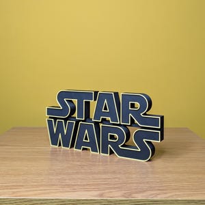 Peut inclure: Logo Star Wars noir et or, pos&eacute; sur une surface en bois contre un fond jaune. Les lettres noires sont bord&eacute;es d'or, cr&eacute;ant un effet 3D. Le logo est un objet d&eacute;coratif.