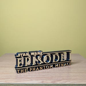 May include: A black and gold 3D printed sign that reads "Star Wars Episode I The Phantom Menace".