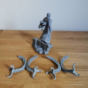May include: A gray plastic figure of a person wearing a long coat and holding a weapon. The figure is standing on a gray plastic base with a detailed design. Four gray plastic tentacles are lying on a wooden surface.