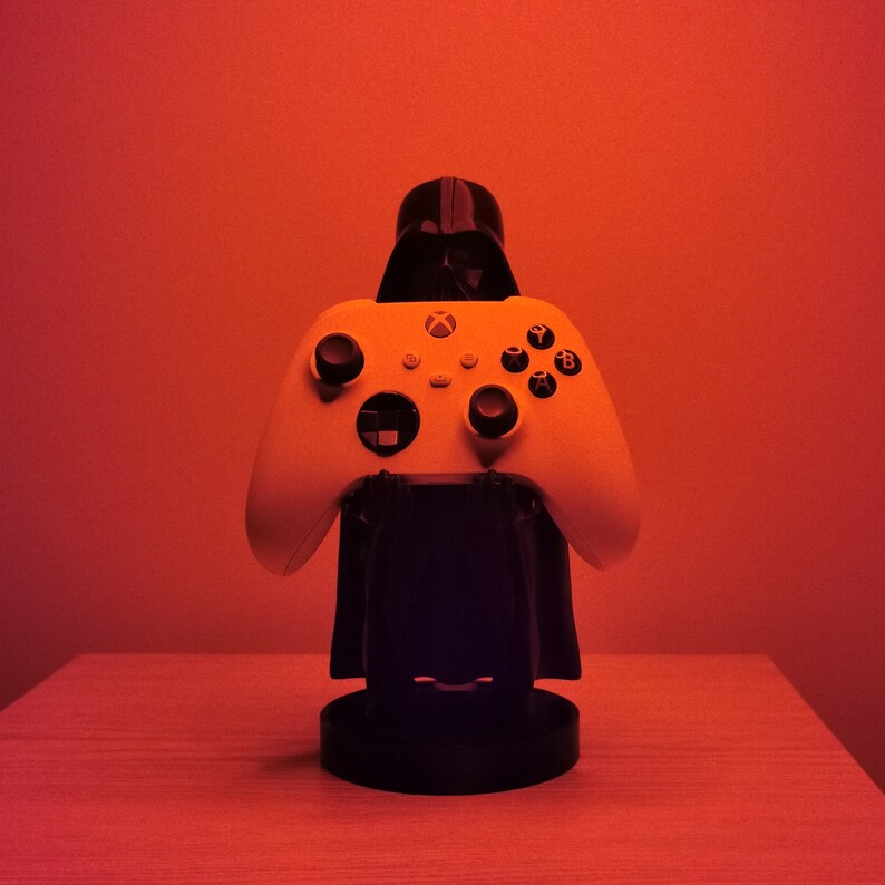 May include: A white Xbox controller with black buttons is held by a black Darth Vader figurine. The controller and figurine are on a black stand.