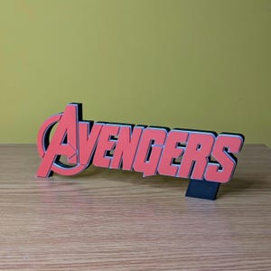 May include: A red and black Avengers logo, a decorative item, stands on a wooden surface. The word "Avengers" is in a bold, blocky font with a red color and a black outline. The logo is a stylized "A" with a red and black color scheme.