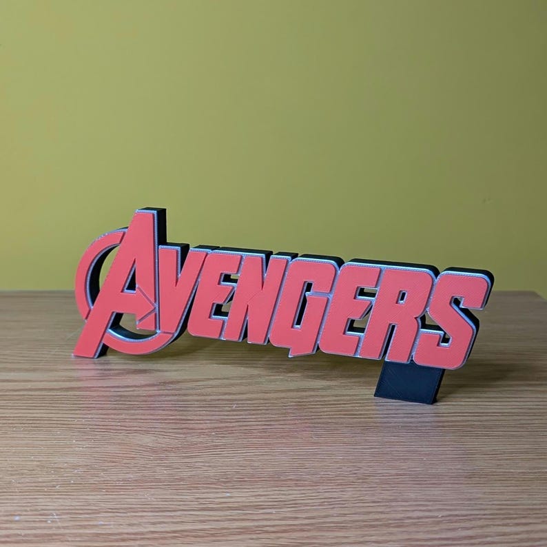 May include: A red and black Avengers logo, a decorative item, stands on a wooden surface. The word "Avengers" is in a bold, blocky font with a red color and a black outline. The logo is a stylized "A" with a red and black color scheme.