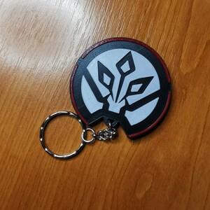 May include: A black and white keychain with a red border. The keychain features a stylised design of a geometric shape with a white centre.