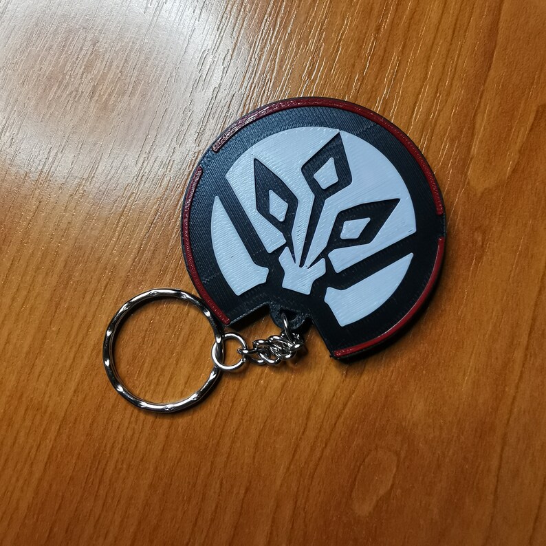 May include: A black and white keychain with a red border. The keychain features a stylised design of a geometric shape with a white centre.