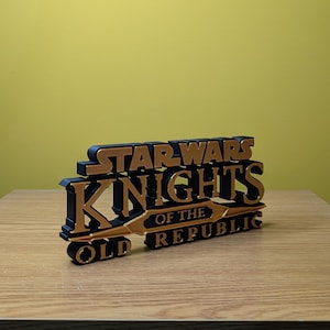 Może przedstawiać: Tr&oacute;jwymiarowy, wydrukowany model logo gry Star Wars: Knights of the Old Republic. Logo jest czarne z złotymi akcentami i zawiera tytuł gry.