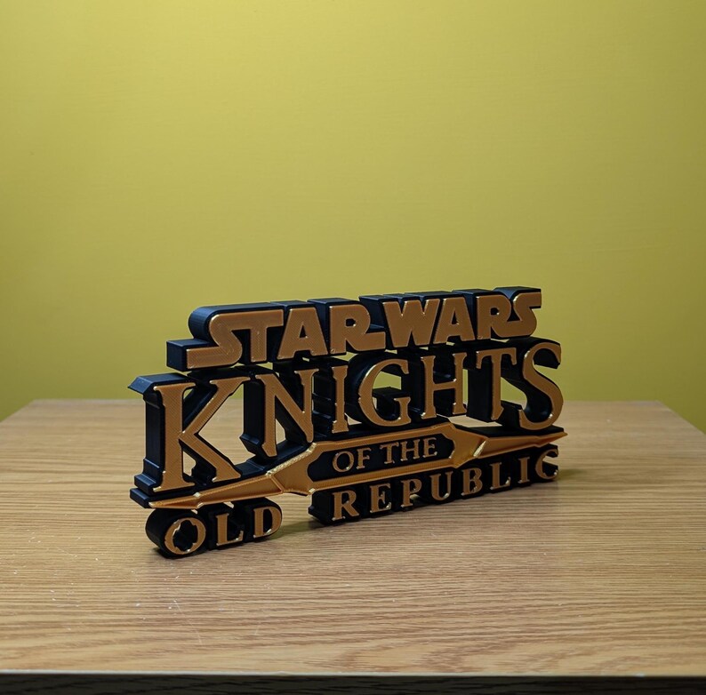 Może przedstawiać: Tr&oacute;jwymiarowy, wydrukowany model logo gry Star Wars: Knights of the Old Republic. Logo jest czarne z złotymi akcentami i zawiera tytuł gry.
