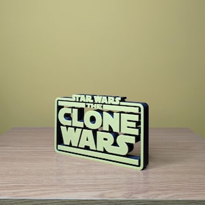 May include: A 3D printed sign with the text "Star Wars The Clone Wars" in yellow letters with a black outline. The sign is on a wooden surface.