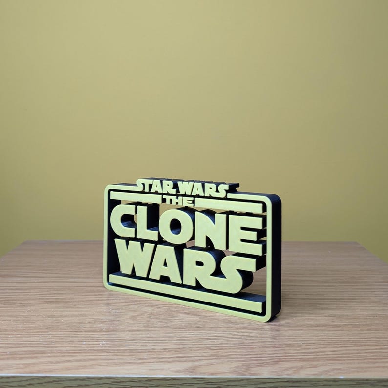 May include: A 3D printed sign with the text "Star Wars The Clone Wars" in yellow letters with a black outline. The sign is on a wooden surface.