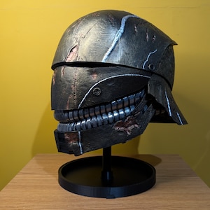 May include: A weathered, dark gray helmet with a silver jaw piece and a stand. The helmet has a distressed look with visible scratches and wear.