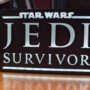 以下が含まれることがあります： 「STAR WARS JEDI SURVIVOR」と白文字で書かれた黒い看板です。