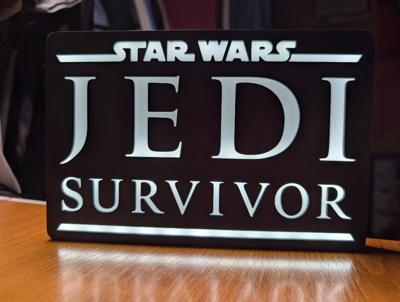 以下が含まれることがあります： 「STAR WARS JEDI SURVIVOR」と白文字で書かれた黒い看板です。