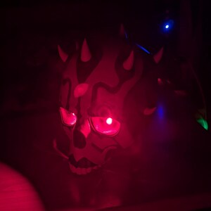 May include: A red and black mask with glowing red eyes and horns. The mask has a skull-like design with intricate patterns and is illuminated by red light, creating a dramatic effect. The background is dark.