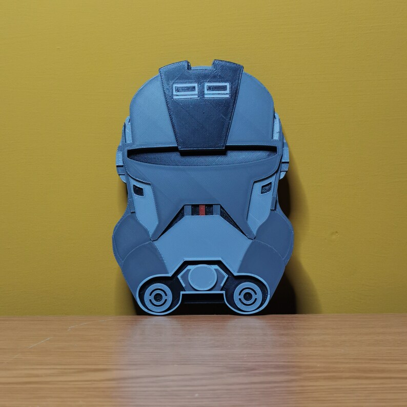 Pu&ograve; includere: Un casco grigio e nero stampato in 3D con una striscia rossa sul viso. Il casco ha una visiera e due piccoli cerchi neri sulla parte superiore.