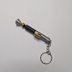 May include: A silver and black lightsaber keychain with a gold accent. The keychain has a silver ring and chain. The lightsaber has a ribbed black handle and a silver top. The keychain is a miniature replica of a lightsaber.