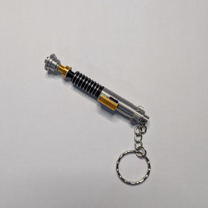 May include: A silver and black lightsaber keychain with a gold accent. The keychain has a silver ring and chain. The lightsaber has a ribbed black handle and a silver top. The keychain is a miniature replica of a lightsaber.