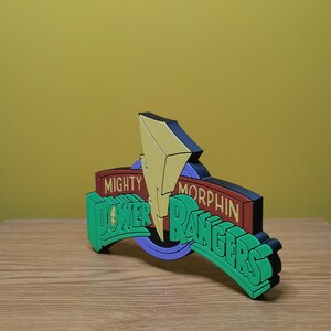 Pu&ograve; includere: Logo dei Power Rangers tridimensionale, nei colori verde, rosso, giallo e nero. Il logo presenta le parole "Mighty Morphin Power Rangers" con un fulmine. Il logo &egrave; su una superficie di legno.