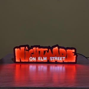 May include: A glowing red and orange sign with the text "A Nightmare on Elm Street." The sign has a black border and is illuminated from within. The sign is resting on a wooden surface.