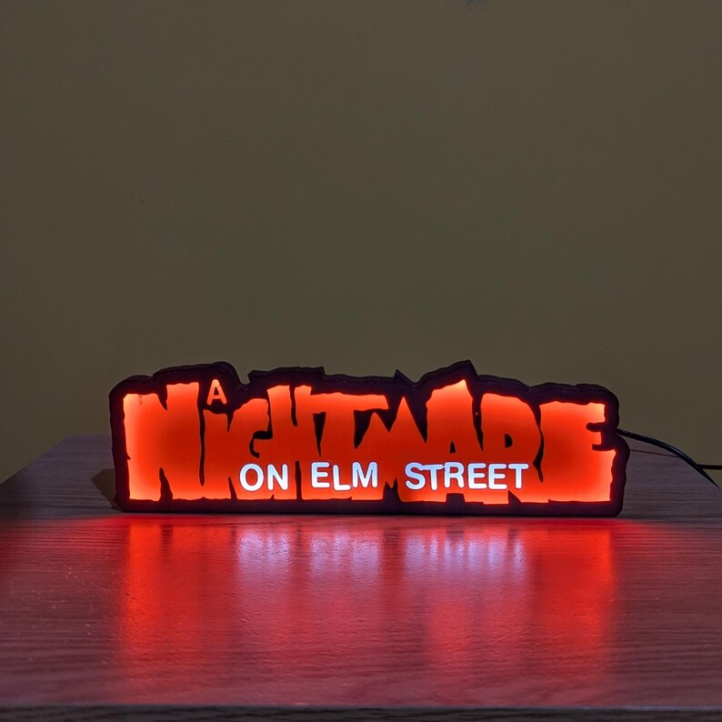 May include: A glowing red and orange sign with the text "A Nightmare on Elm Street." The sign has a black border and is illuminated from within. The sign is resting on a wooden surface.