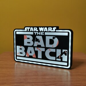 May include: A black and white sign with white LED lights around the perimeter. The sign reads "Star Wars The Bad Batch" in white letters with a red and white splatter pattern behind the letters.