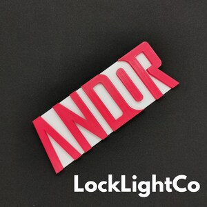 Op de afbeelding: Een rechthoekig, rood en wit bord met het woord "ANDOR" in vette, rode letters. Het bord is schuin, waarbij de letters verhoogd lijken. De tekst "LockLightCo" is in het wit onderaan gedrukt.