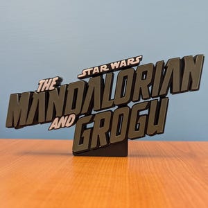 Op de afbeelding: Een zwart en grijs bord met de tekst "The Mandalorian and Grogu" en "Star Wars" erboven. Het bord is 3D en staat op een kleine zwarte basis. Een decoratief item.